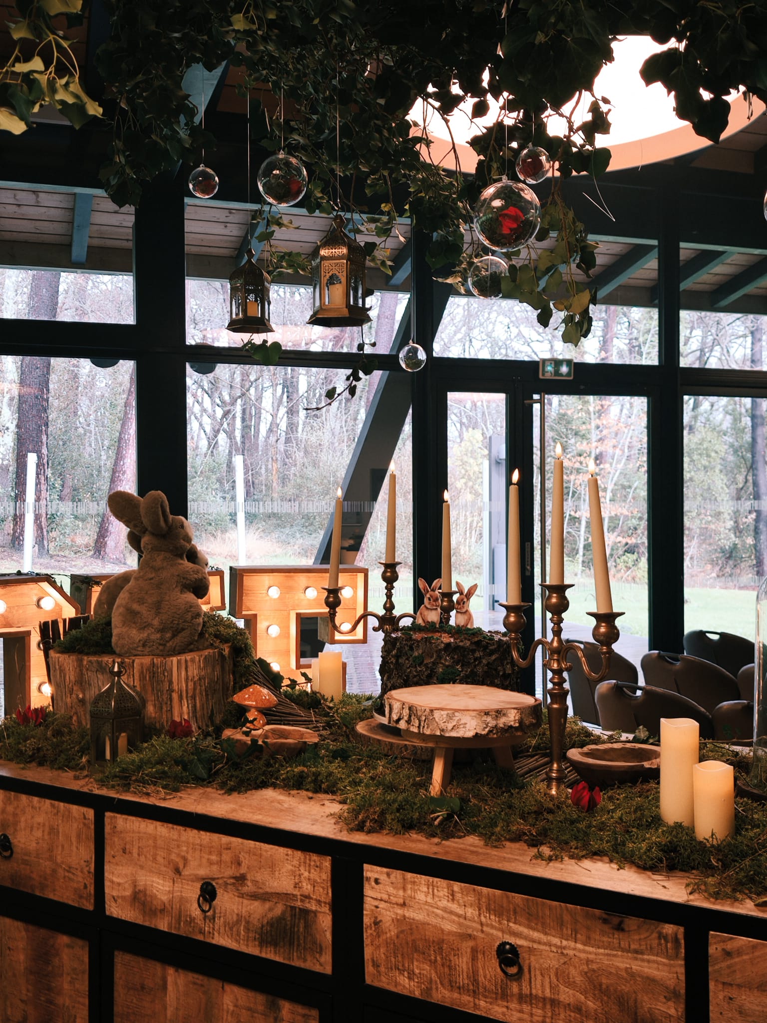 La table centrale de la réception était une véritable installation artistique à elle seule. Recouvert d'un épais tapis de mousse verte naturelle, le plateau accueillait une composition d'objets soigneusement sélectionnés pour raconter l'histoire de la forêt : champignons en céramique orange et rouge à pois blancs, rondelles de bois brut servant de plateaux ou de dessous de verre, chandeliers en laiton doré vieillis portant des bougies pilier ivoire, et — au centre, pièce maîtresse — deux lapins en peluche enlacés, posés dans un tronc creux comme s'ils s'y étaient installés naturellement. Cette table n'était pas simplement fonctionnelle : elle était narrative. Chaque détail participait à construire un récit cohérent, celui d'un amour solide et tendre, célébré dans l'intimité d'une forêt magique.